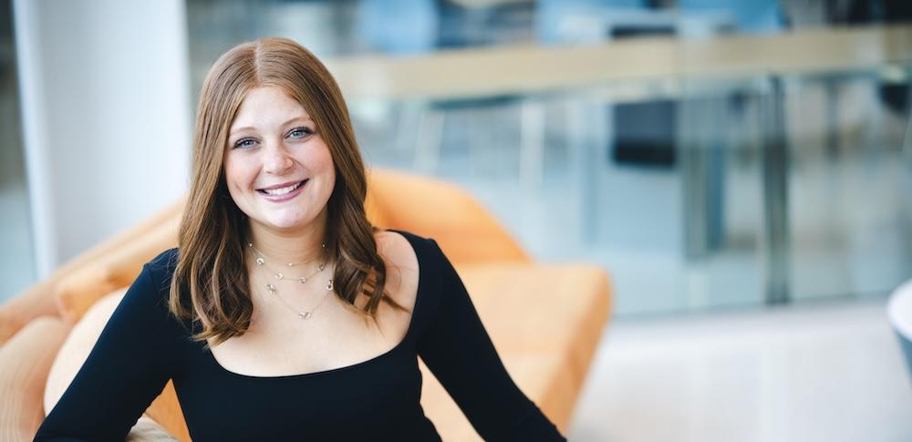 Julia Klayman sits on a sofa and smiles at the camera. She wears a black long sleeved shirt.